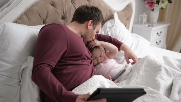 El joven padre y la pequeña hija están usando aparatos acostados en la cama. Hombre guapo está besando y abrazando a su niño inteligente . — Vídeos de Stock