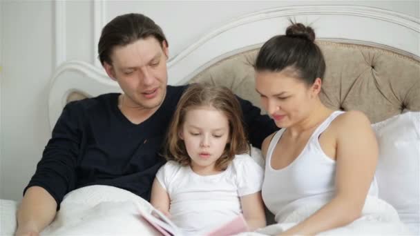 Linda menina está lendo em voz alta o livro deitado na cama com seus pais. Pai bonito está beijando seu filho inteligente . — Vídeo de Stock