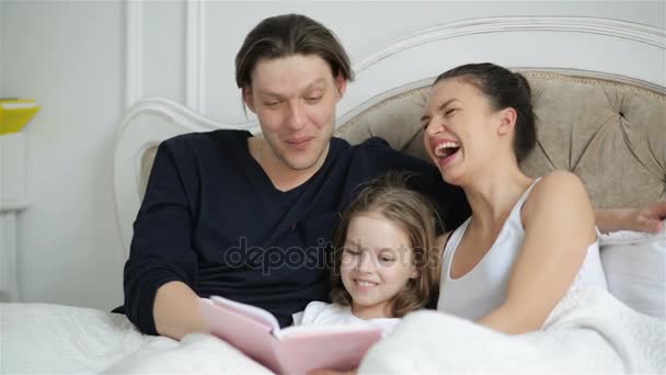 Roztomilá malá dcera je čtení nahlas knihu v cizí jazyk ležící na posteli, její rodiče se smějí v některých cizích slov. Vtipné momenty studia. — Stock video