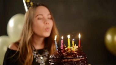 Sevimli genç kız uzun saçlı bir çikolatalı kek ayakta altın ve beyaz hava balonları siyah arka plan üzerinde mum üfleme olduğunu. Doğum kız ve tatlı tatlı.