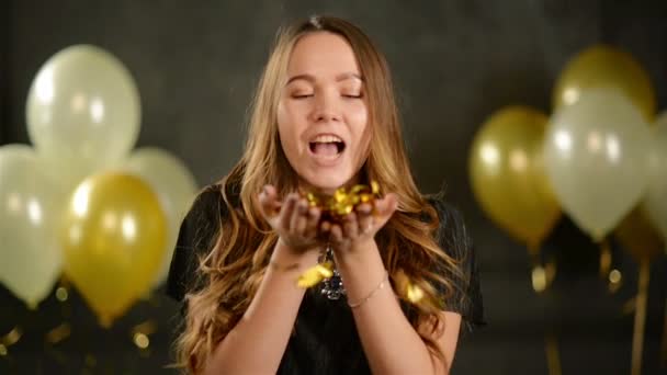 Primer plano Retrato de la muchacha del cumpleaños con puñado de Confetti de oro. Mujer joven sonriente está soplando en sus manos disfrutando de la fiesta. Fondo negro con globos de aire — Vídeos de Stock