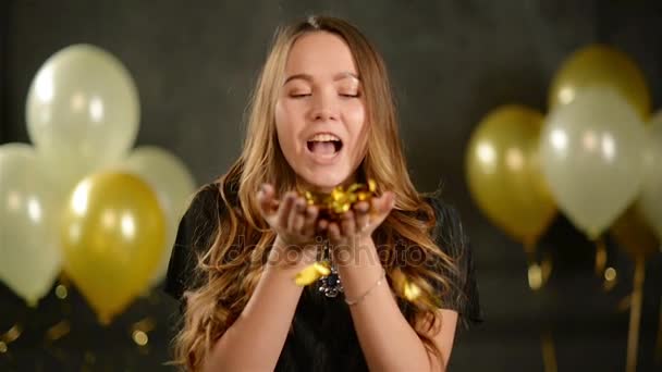 Vértes portré szülinapos maroknyi arany konfetti. Mosolygó fiatal nő az ő kezét élvezi fél Blowing. Fekete háttér levegő léggömbök