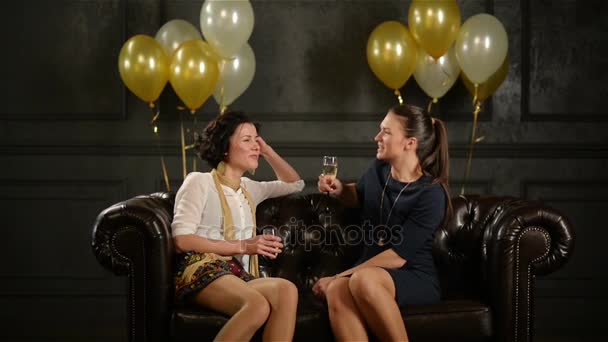 Due Pretty Lady sono sedute sul divano marrone con bicchieri di champagne e parlano di qualcosa di divertente durante una festa. Muro nero con palloncini d'aria dorati e bianchi sullo sfondo . — Video Stock