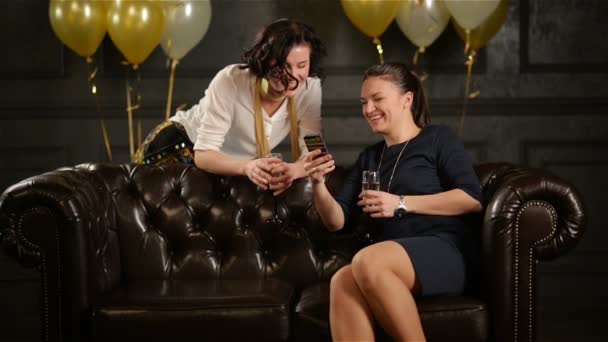 Mulheres sorridentes com copos de champanhe nas mãos estão usando Smartphone durante a celebração. Lady with Short Curly Hair is Standing next to Brown Sofa, Another One está sentado sobre ele . — Vídeo de Stock