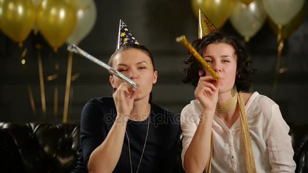 Duas mulheres brincalhões estão soprando em apitos animadores durante uma celebração. Closeup Retrato de morenas engraçadas em tampas de aniversário em fundo preto com balões de ar . — Vídeo de Stock