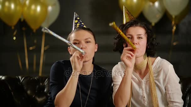 Dos mujeres juguetonas están sonando en silbatos de porrista durante una celebración. Primer plano retrato de morenas divertidas en gorras de cumpleaños sobre fondo negro con globos de aire . — Vídeo de stock