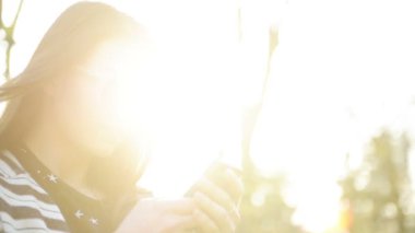 Güneş ışınları zevk mobil aygıt kullanarak ve sıcak gün açık havada güneş gözlüğü ve uzun saç sevimli kadındır. Parkta Smartphone ile esmer gülümseyen portresi.