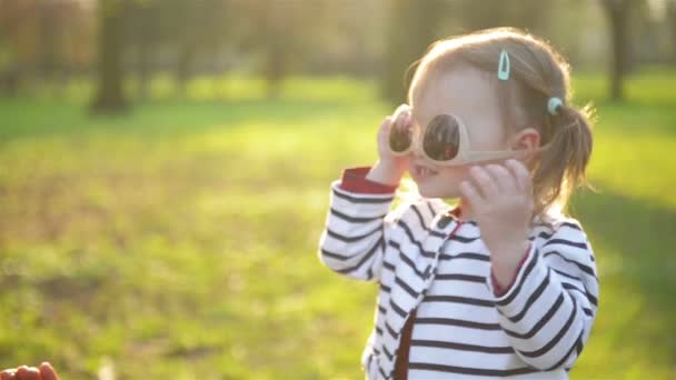 Little Daughter está tratando de usar gafas de sol de madres, Young Mom Hepls Her. Morena de moda y chica bonita con dos colas de caballo están disfrutando del clima soleado en el parque en primavera . — Vídeos de Stock