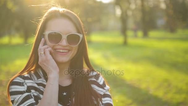 Усміхаючись брюнетка з довгим волоссям і Fashional сонцезахисних окулярів є Talking мобільного телефону під час сонячний день весни в парку. Портрет красива жінка з смартфон на відкритому повітрі. — стокове відео