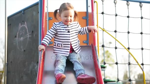 Funny Cute Girl con dos colas de caballo está jugando en la diapositiva roja. Alegre niña con chaqueta a rayas divirtiéndose en el parque infantil . — Vídeos de Stock