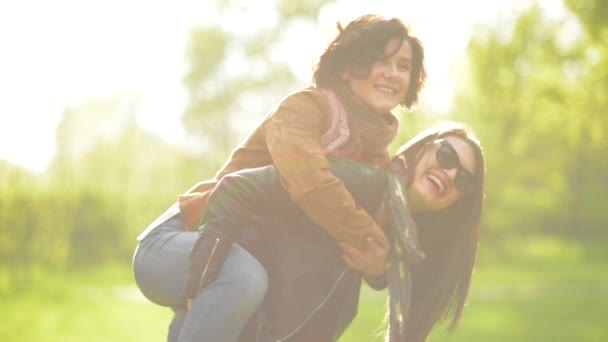 Happy Female Friends se está divirtiendo afuera disfrutando de la luz del sol en el parque. Mujer joven con el pelo largo y gafas de sol está llevando otro en sus hombros en fondo de hierba verde . — Vídeos de Stock