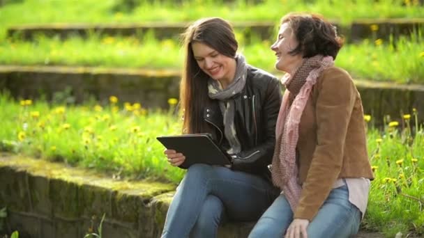 Las mujeres jóvenes con sonrisas dentadas están usando tabletas en las manos. Atractivas morenas se están divirtiendo junto con un dispositivo electrónico en el parque durante el soleado día de primavera al aire libre . — Vídeo de stock