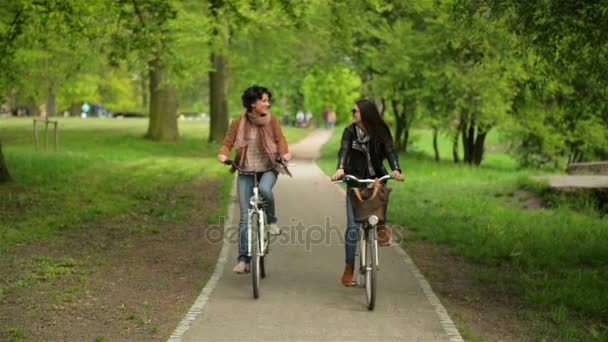 Dos mujeres de cabello oscuro están montando por el parque de la ciudad. Hermosas morenas con bicicletas disfrutando del clima cálido y soleado al aire libre . — Vídeos de Stock