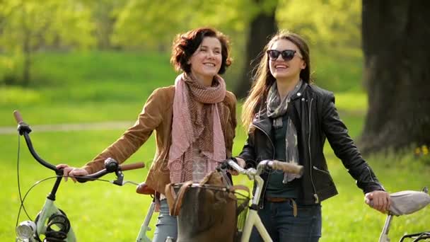 Atractivas morenas con bicicletas pasan tiempo juntas en el parque. Dos mujeres jóvenes activas conversando al aire libre sosteniendo sus bicicletas . — Vídeos de Stock