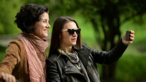 Dos mujeres jóvenes activas con bicicletas están haciendo selfie usando el teléfono móvil. Retrato al aire libre de morenas lindas sonrientes con bicicletas Thier en el parque . — Vídeo de stock