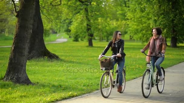 Las amigas despreocupadas están montando juntas y sonriendo. Dos mujeres bonitas con bicicletas se divierten afuera en el parque . — Vídeo de stock