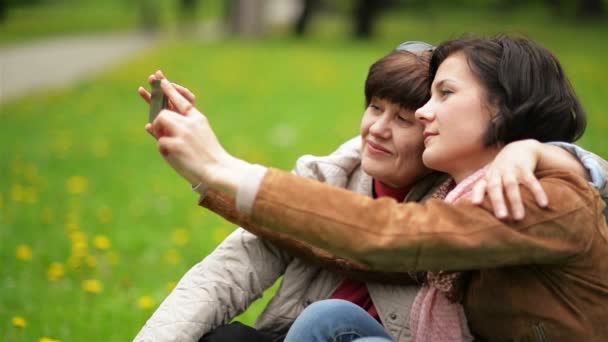 Olgun kızı ve onun annesi kullanma Smartphone sırasında piknik Park vardır. Selfie açık havada alarak iki gülen esmer kadın vardır. — Stok video