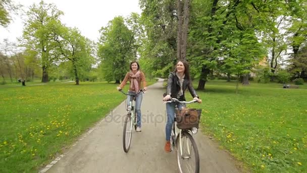 Vista frontal de dos amigas montando en bicicleta en el parque de la ciudad en primavera. Cámara en movimiento . — Vídeos de Stock