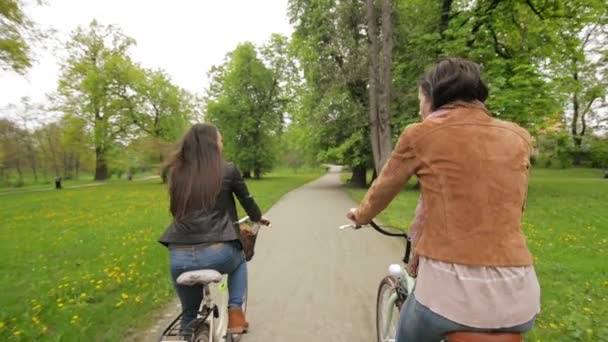 Vista trasera de dos morenas en chaquetas y pantalones vaqueros montando en bicicletas en el parque en primavera. Cámara en movimiento . — Vídeos de Stock