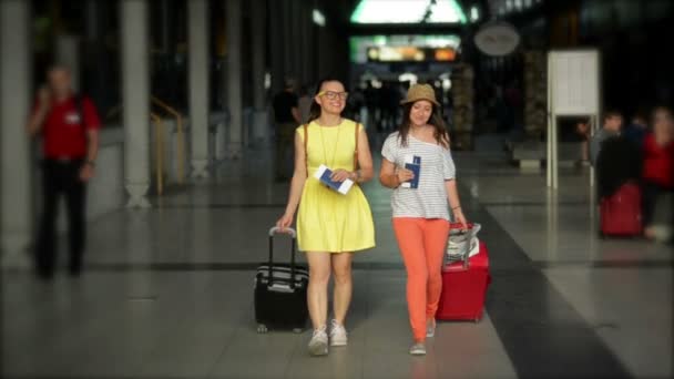 Две улыбающиеся девушки катаются большими сумками в аэропорту. Красотка в жёлтом платье и очках путешествует со своей подругой в полосатой рубашке и шляпе . — стоковое видео