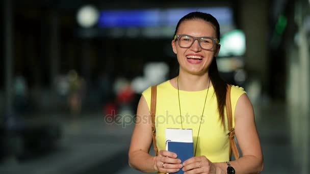 微笑的年轻女士是抱着她护照和机票在机场的手中。漂亮的女孩在眼镜和黄色的连衣裙，等待飞机的画像。背景模糊的记分牌. — 图库视频影像