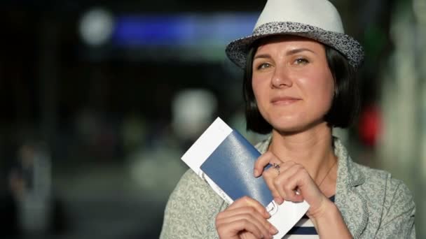 Young Woman with Short Dark Hair and Summer Hat Holding Her Passport and Ticket is Looking at the Camera and Smiling at the Airport. Blurred Scoreboard on the Background. — Stock Video