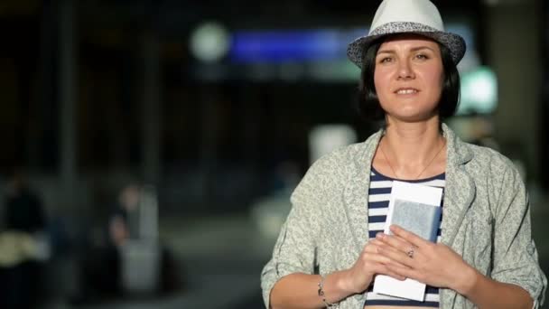 Porträt einer jungen Brünetten mit kurzen Haaren, Sommermütze und gestreiftem Kleid, die ihren Pass und ihr Ticket am Flughafen vor der Kamera zeigt. verschwommene Anzeigetafel auf dem Hintergrund. — Stockvideo