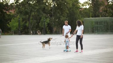 Kentsel şehir meydanında mutlu aile portresi. Yakışıklı erkek ile küçük köpek, genç kadın tutan Her kızı el, kız elbise Rides bir Scooter üzerinde gider.