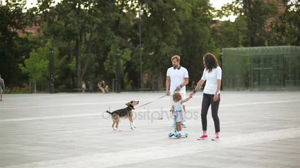 Portrait urbain de la famille heureuse sur la place de la ville. bel homme va avec petit chien, jeune femme tient sa main filles, fille en robe monte sur un scooter . — Video