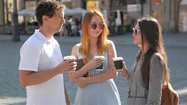 Dos chicas en gafas de sol y vestidos de moda están hablando con un chico guapo con camisa blanca y pantalones vaqueros en la calle. Tres amigos tomando café al aire libre durante el día cálido de verano . — Vídeos de Stock