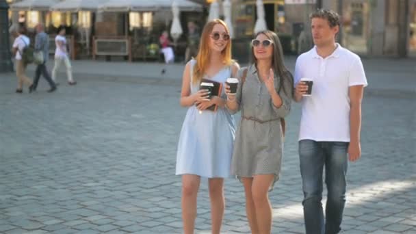 Tres amigos tomando café y caminando por la ciudad durante el día cálido de verano. Dos chicas con gafas de sol y vestidos cortos, chico guapo con camisa blanca y pantalones vaqueros sonriendo y hablando con las mujeres . — Vídeos de Stock