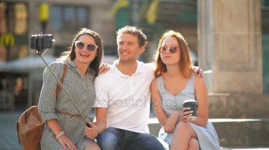 Önden Görünüm Smartphone açık havada güneşli yaz gün boyunca şehir meydanında kullanarak Selfie alarak üç mutlu arkadaş. İki inanılmaz kız ve bir erkek arıyorum kameraya gülümseyen.