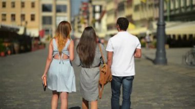 Arkadan Görünüm üç arkadaş kahve içme ve şehirde yürüyüş. Güneş gözlüğü ve kısa elbiseler ve yakışıklı çocuk beyaz gömlek ve birlikte vakit kot giyen iki kız.