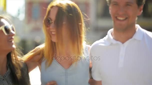 Tres amigos felices se reúnen en la acera de una calle de una gran ciudad. Dos niñas y un niño abrazándose en el fondo urbano al aire libre durante el cálido día de verano . — Vídeo de stock