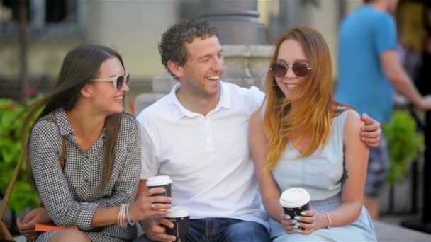 Ritratto di tre amici che bevono caffè seduti sulla panchina all'aperto in città durante la calda giornata estiva. Ragazzo sta abbracciando ragazza in abito blu chiaro. Un'altra donna ha lunghi capelli scuri e occhiali da sole . — Video Stock