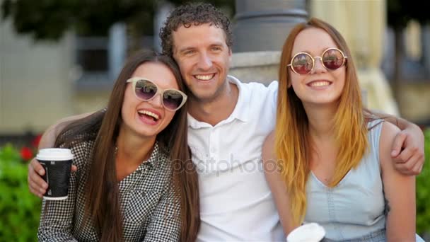 Όμορφος άνδρας είναι το αγκάλιασμα δύο όμορφα κορίτσια σε μοντέρνα γυαλιά ηλίου. Τρεις φίλους χαμογελώντας θέτοντας κοιτάζοντας την κάμερα σε εξωτερικούς χώρους κατά τη διάρκεια της θερινής ώρας. — Αρχείο Βίντεο