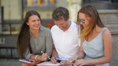 Üç öğrenci açık havada çalışmak ve birlikte sıcak güneşli gün boyunca şehirde gülüyor. İki kız ve bir erkek notlar, kalem ve Tablet Prepearing sınavı.