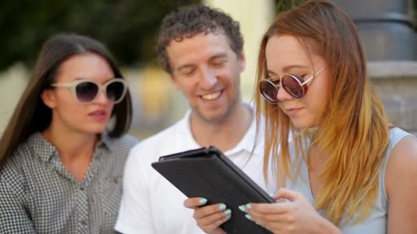 Grupo de estudiantes con Tablet Hanging out. Chica en vestido azul claro está leyendo algo, su amiga y novio escuchar sentado al aire libre durante el cálido día soleado en la ciudad . — Vídeos de Stock