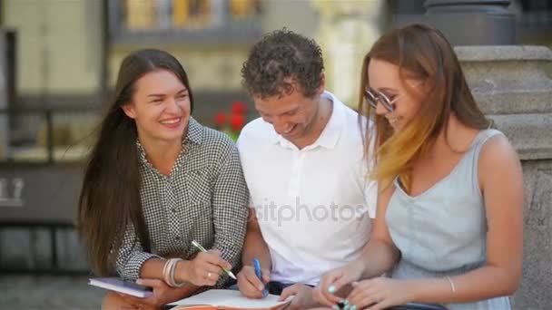 Tre studenti che studiano all'aperto e ridono insieme durante la calda giornata di sole in città. Due ragazze e un ragazzo con note, penne e tablet Prepearing per l'esame . — Video Stock