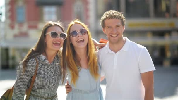 Zwei süße Frauen mit langen Haaren, Sonnenbrille und kurzen Kleidern amüsieren sich zusammen mit einem gutaussehenden Mann in weißem Hemd in der Altstadt an einem sonnigen Sommertag. — Stockvideo