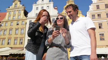 Eski Şehir Meydanı üzerinde duran gülümseyen genç insanlar. Smartphone kullanarak ve bir şey onun için gösterilen bir ekran dokunarak şirin esmer.
