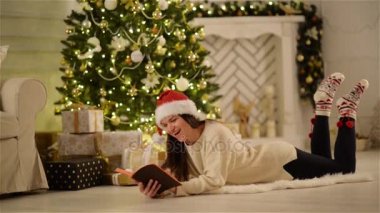 Uzun saçlı bir açılan kitap Her el ve gülen Xmas ağacı arka planda tutan ne? Noel Baba şapkası komik hikayeler okuma ile oldukça esmer yere Noel döşeme ev dekore edilmiş.