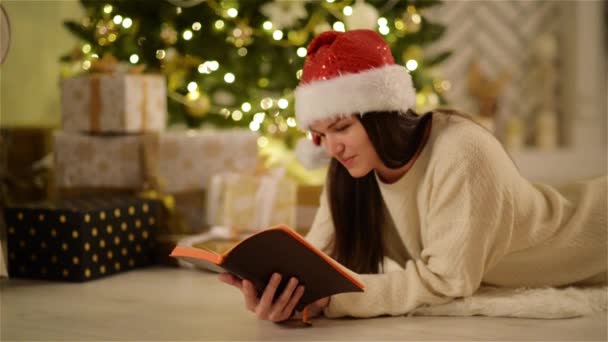 Mujer feliz asombrada en Santa Sombrero con el pelo largo acostado en el suelo y disfrutando de la lectura. Retrato de cerca de una niña sonriente sosteniendo un libro abierto en las manos junto al árbol de Navidad y cajas con regalos . — Vídeo de stock