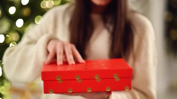 Onherkenbaar Brunette Opening kerstcadeau, binnenshuis. Uitpakken van het rode vak met heden. Wazig Kerstverlichting op achtergrond. — Stockvideo