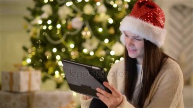 Noel ağacı arka plan ellerinde Tablet ile şirin esmerle closeup portresi. Noel Baba gülümseyerek ve Noel kutlamaları sırasında Gadget'ı kullanarak şapkalı duygusal kız görünümünü.
