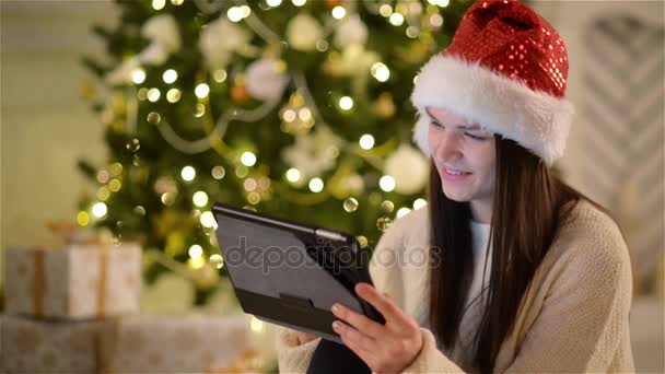 Portret zbliżenie ładny brunetka z tabletu w jej ręce na tle choinki. Zobacz dziewczynka emocjonalność w Santa Hat uśmiechając się i korzystając z gadżetu podczas obchodów Święta. — Wideo stockowe