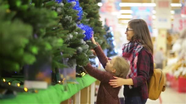 スーパーで母と子を選択するクリスマス ツリー。若く美しい母と娘を買うスーパー マーケットの棚、メリー クリスマスと幸せな新年の近くの装飾とクリスマス ツリー — ストック動画