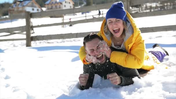 Joyous Girl in Yellow Coat is rubbing the Face of Her Boyfriend by Snow. Шутка о влюбленной девушке во время зимних каникул в горах . — стоковое видео
