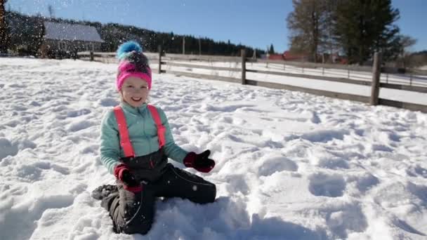 Joyous Little Girl se está divirtiendo en la nieve sentada en el suelo y lanzando copos de nieve al aire. Día soleado de invierno. Concepto de la infancia feliz . — Vídeo de stock