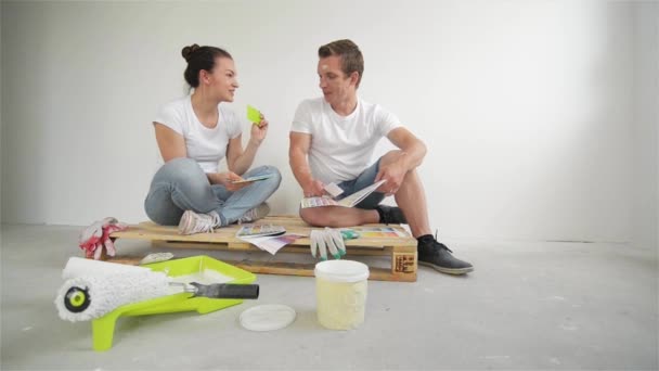 Glada unga par planering designprojekt för deras lägenhet som sitter på golvet med palett av färger på vit vägg bakgrund. — Stockvideo